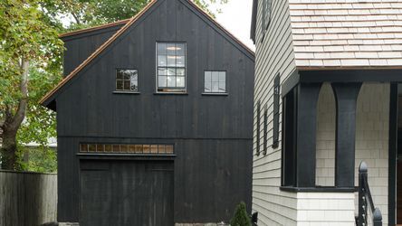 A dark-colored, two-story detached garage with a pitched roof and large doors stands next to a house with light siding. It features vertical wood paneling, a row of small windows above the garage door, and several trees surrounding the structures.